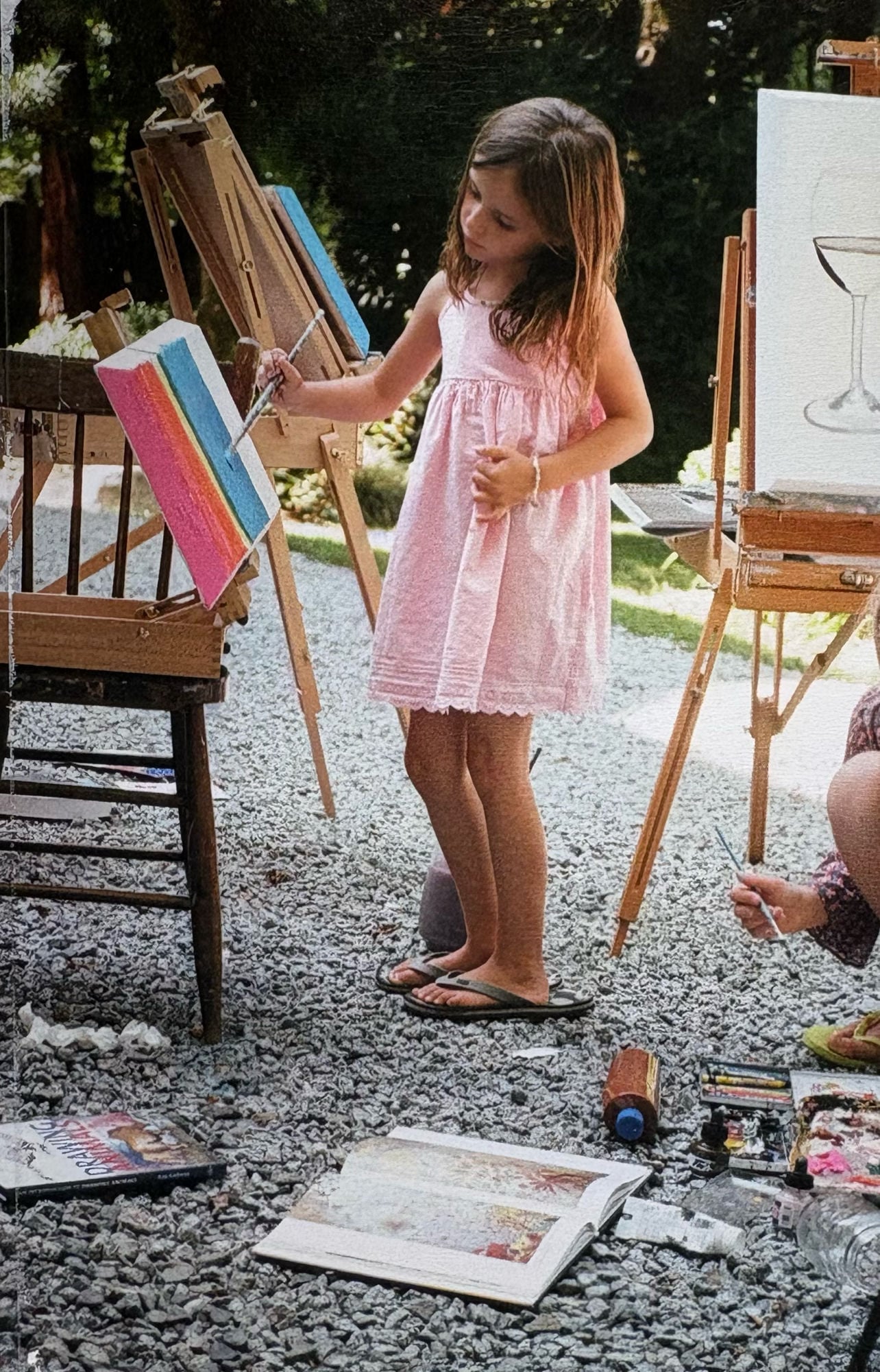 Ella as a young girl painting a rainbow on a small canvas at an outdoor easel